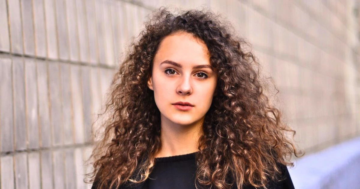 Young woman with long, voluminous natural curly hair showing signs of frizz in curly hair standing against a grey wall background wearing a black top